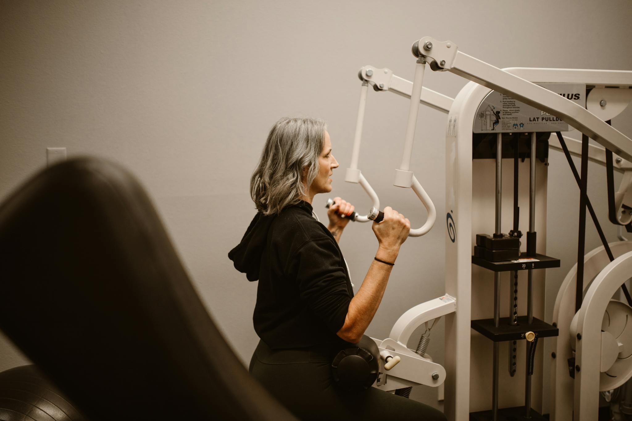 A middle-aged woman with gray hair using a lat pulldown machine for fitness indoors.
