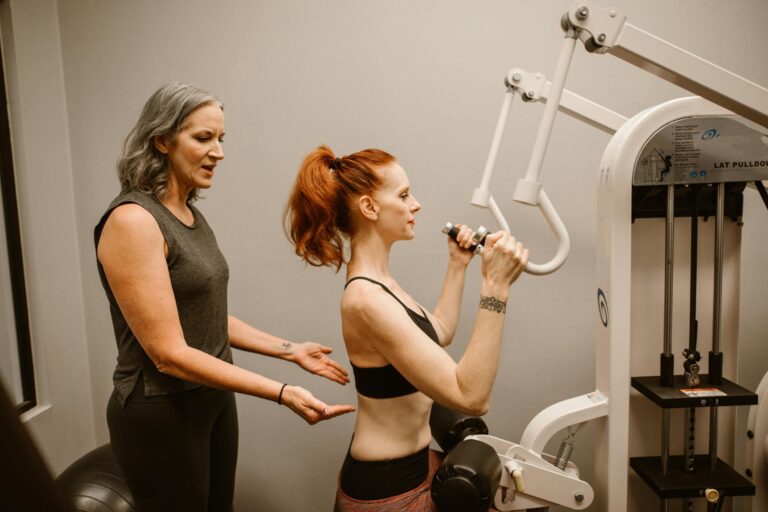 A woman works out on gym equipment with guidance from a trainer indoors.